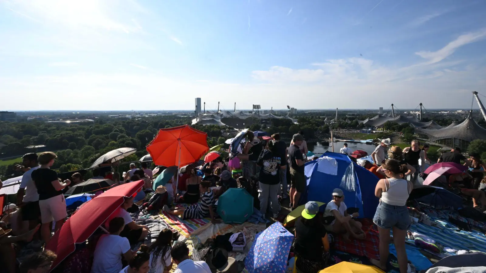 Taylor-Swift-Fans sitzen mit Sonnenschirmen vor der Kulisse des Olympiastadions auf dem Gipfel des Olympiabergs. (Bild: Felix Hörhager/dpa)