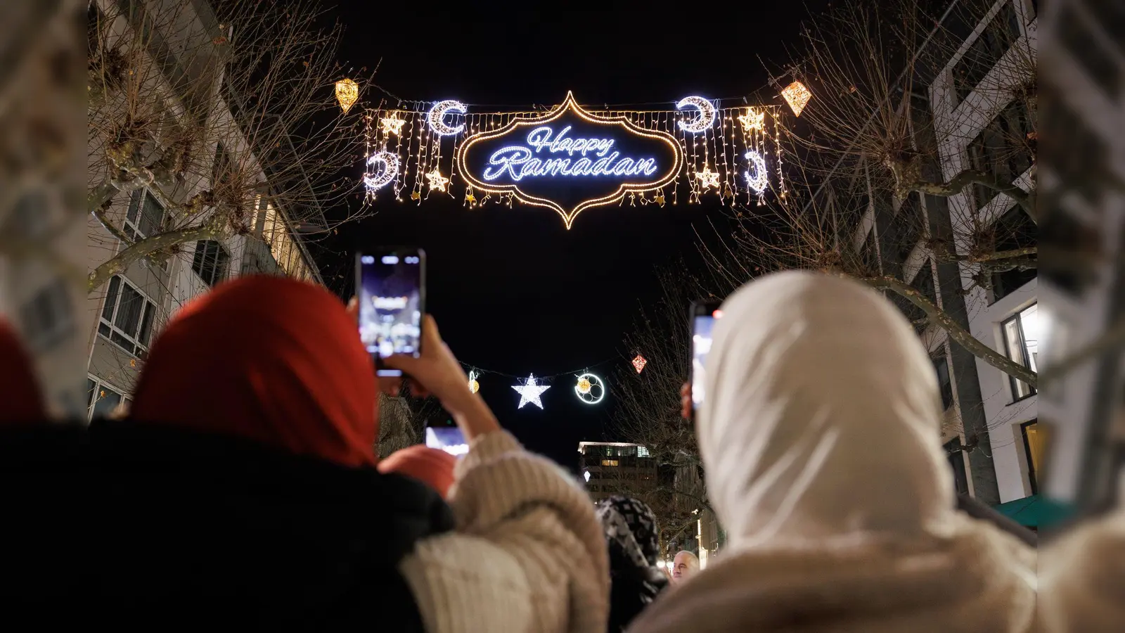 Passanten fotografieren mit ihren Handys die Beleuchtung mit dem Schriftzug „Happy Ramadan” in der Freßgass. Die Beleuchtung wurde 2023 von der Stadtverordnetenversammlung beschlossen und gilt als deutschlandweit einmalig. (Symbolbild: Lando Hass/dpa)