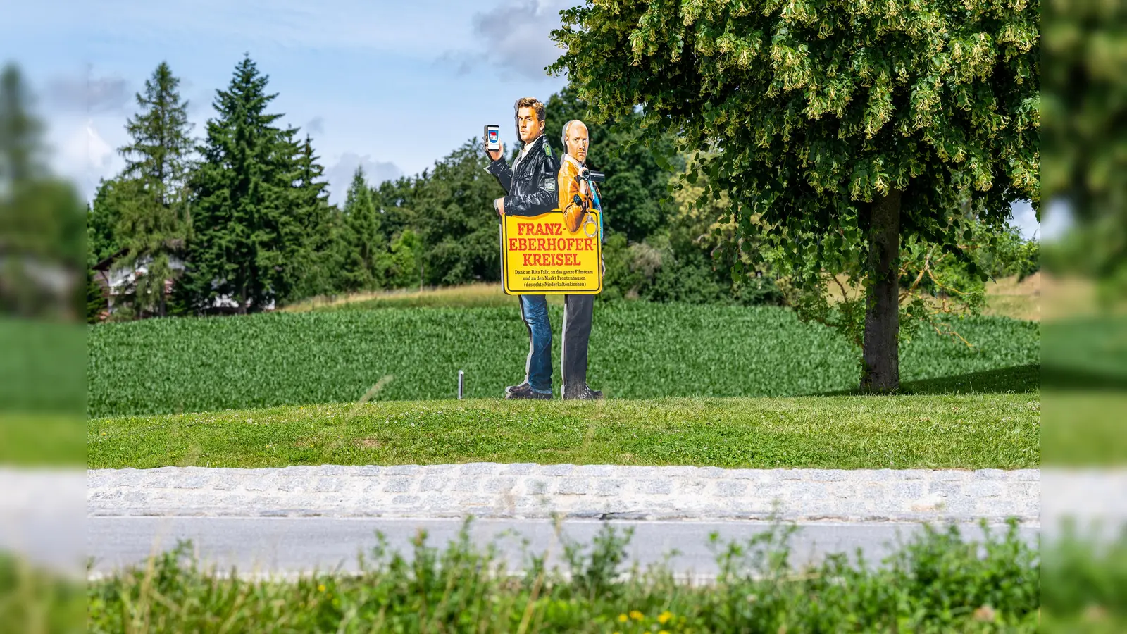 Ein Bild mit den Darstellern der Krimireihe um den Dorfpolizisten Franz Eberhofer und Co-Ermittler Rudi steht auf dem Franz-Eberhofer-Kreisel. Nun wurde die Linde von Unbekannten gefällt. (Archivbild: Armin Weigel/dpa)