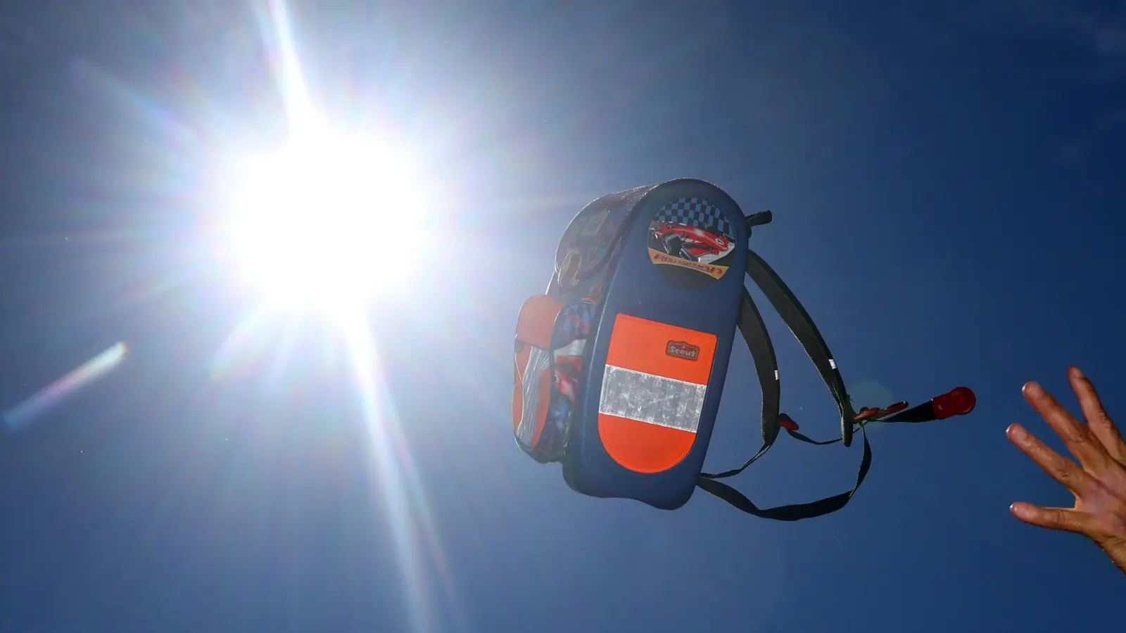 Ein Schulranzen fliegt in der gleißenden Sonne durch die Luft. Hurra, die Schule endet früher! Die Amberger Gymnasien haben angesichts der aktuell heißen Tage den Nachmittagsunterricht gestrichen.  (Symbolbild: Karl-Josef Hildenbrand/dpa)