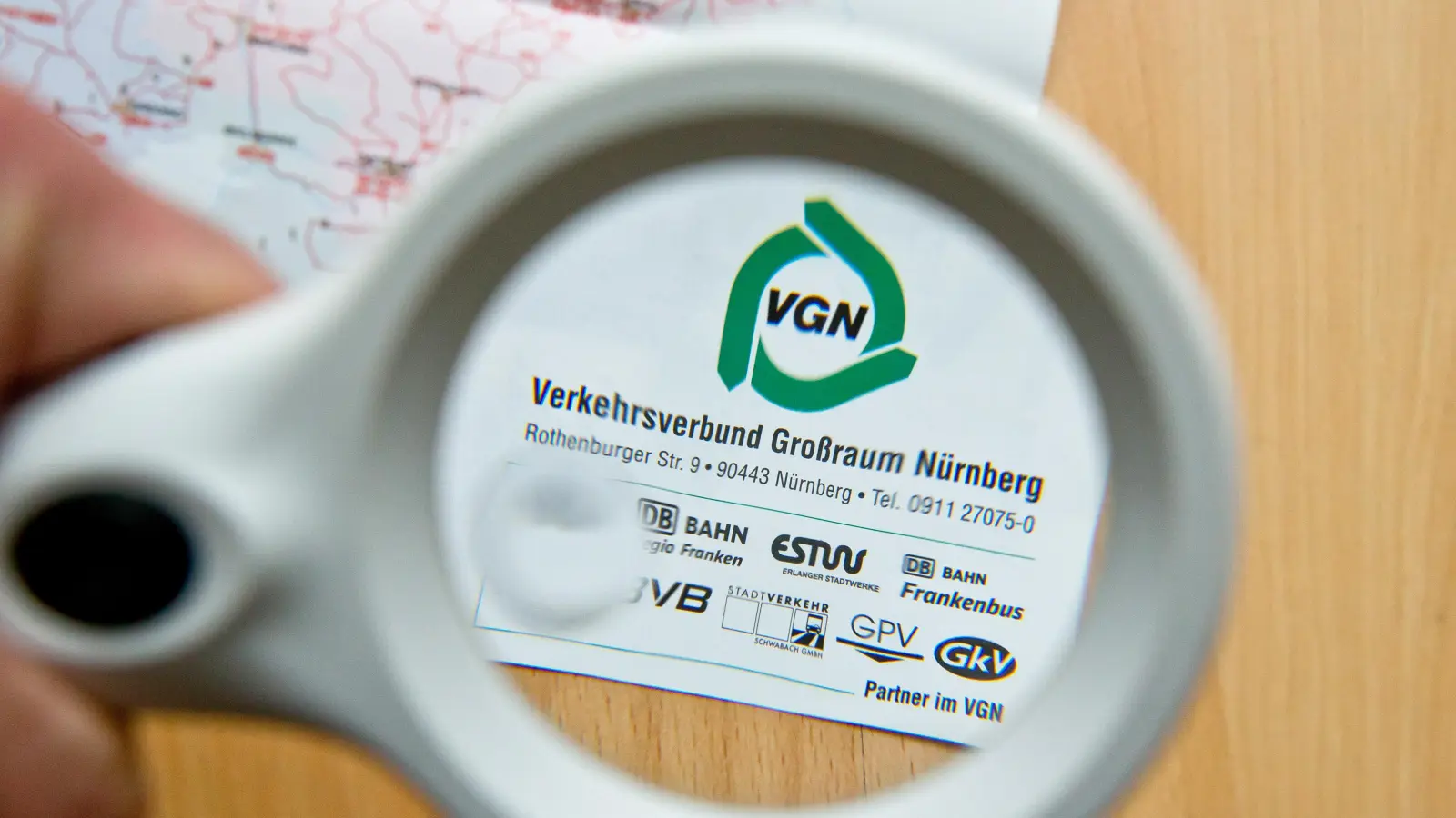 Durch eine Lupe ist auf einer Karte des Verkehrsverbunds Großraum Nürnberg (VGN) das VGN-Logo zu sehen. Dieser begrüßt Neu-Amberger jetzt mit einem Gratis-Ticket. (Symbolbild: Daniel Karmann/dpa)