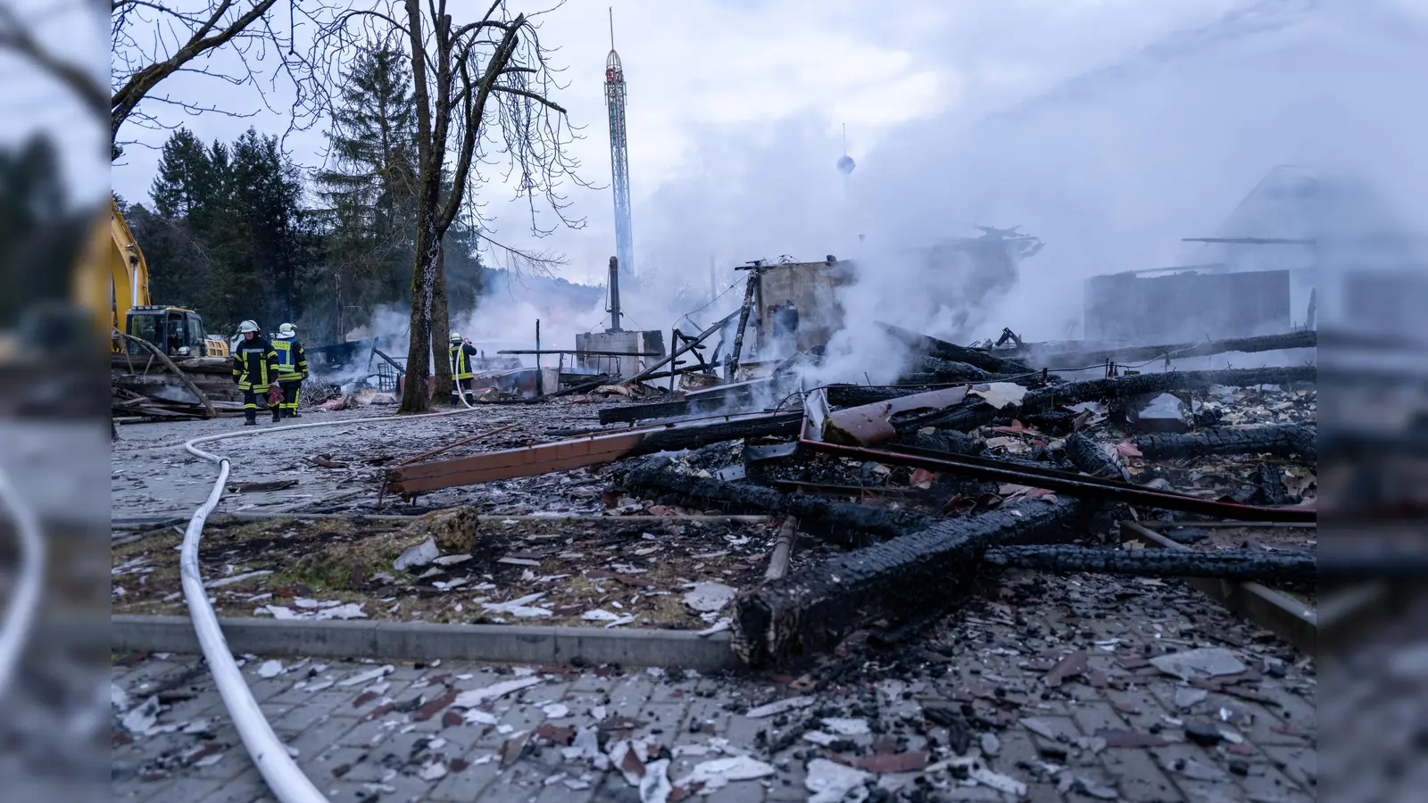 Verkohlte Holzbalken liegen am Boden und Rauch steigt auf, während im Hintergrund der Turm einer Attraktion im Freizeit-Land Geiselwind zu erkennen ist. Im Freizeit-Land Geiselwind ist ein Feuer ausgebrochen. (Bild: Pia Bayer)