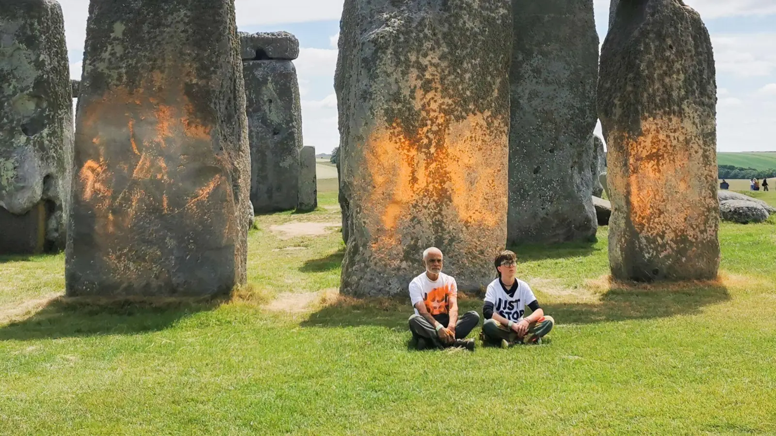 Demonstranten von „Just Stop Oil“ sitzen vor dem Steinmonument Stonehenge, das sie zuvor mit orangener Farbe besprüht haben. (Bild: Just Stop Oil/PA Media/dpa)